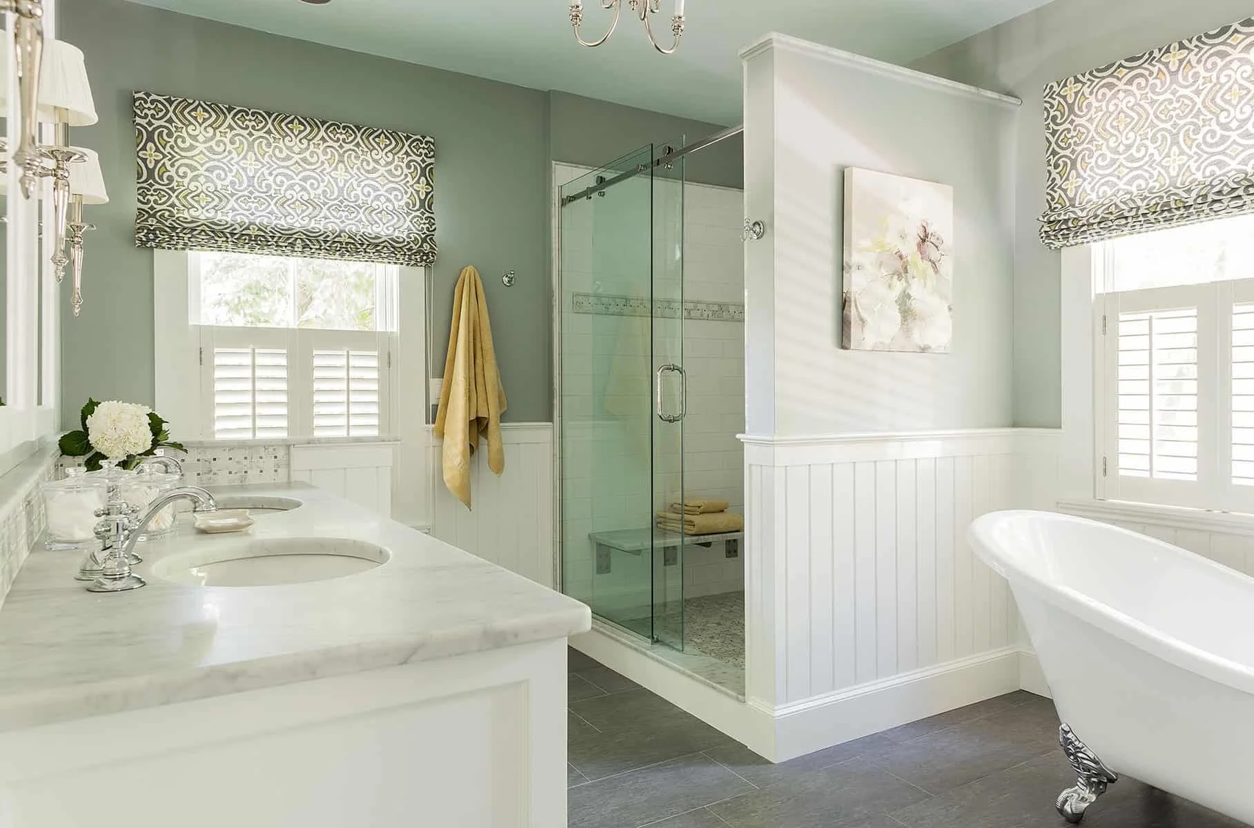 A bright and elegant bathroom featuring a double sink vanity with marble countertop, a glass-enclosed shower, a clawfoot bathtub with decorative silver feet, and windows with patterned Roman shades. The walls are painted light green, and there are wh