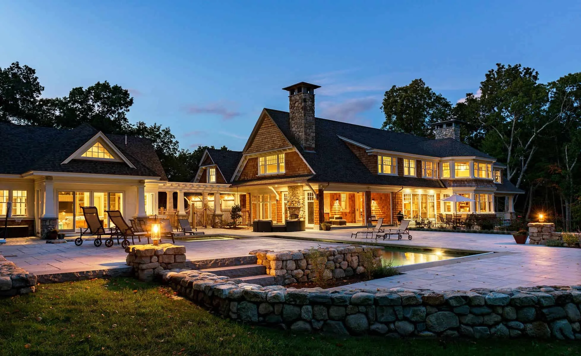 Large shingle-style house with multiple windows and chimneys illuminated at dusk, with a spacious outdoor area featuring lounge chairs, umbrellas, and a firepit, surrounded by trees and a stone patio.