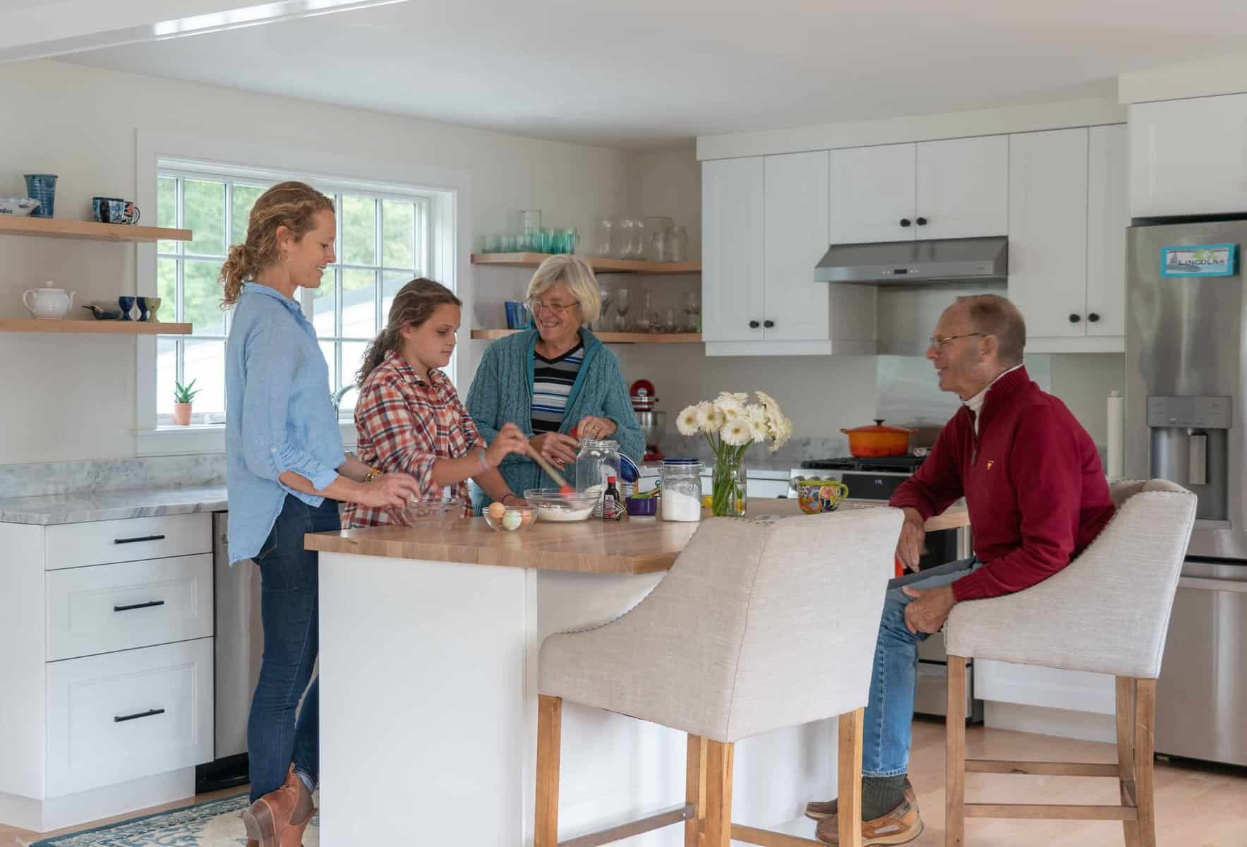 Family gathered in a bright kitchen, with two women, a young girl, and an older man, preparing and sharing a moment together.