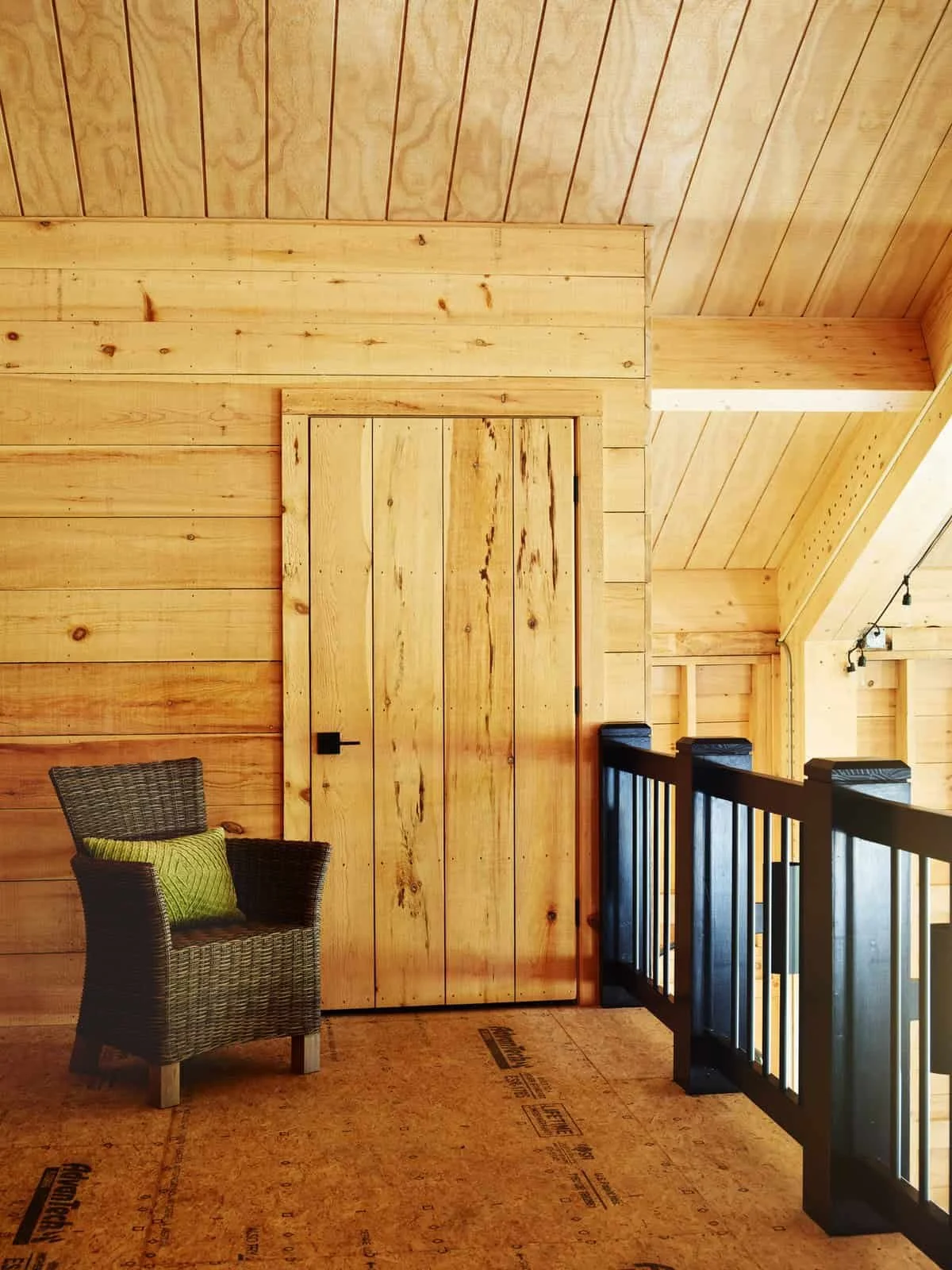 Interior of a wooden cabin with a closed wooden door, a wicker chair with a green pillow, and a black wooden railing, all with natural wood paneling on walls and ceiling.