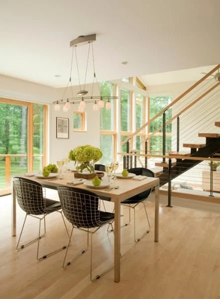 A modern dining area with a wooden table set for four, black wire chairs, a floral centerpiece, and natural light streaming through large windows with a staircase adjacent.