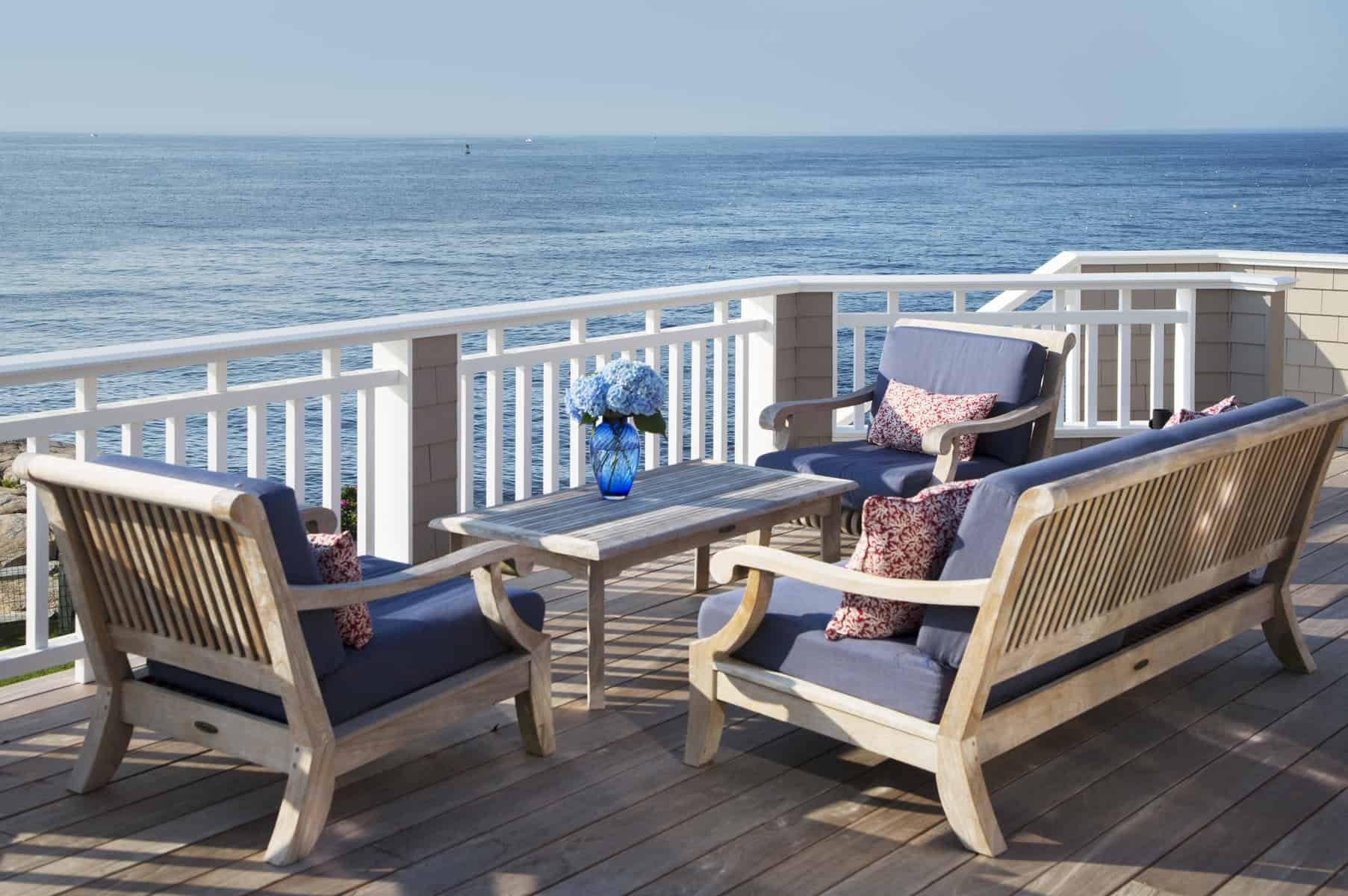 Outdoor porch with wooden furniture and blue cushions, overlooking the ocean with a clear sky.