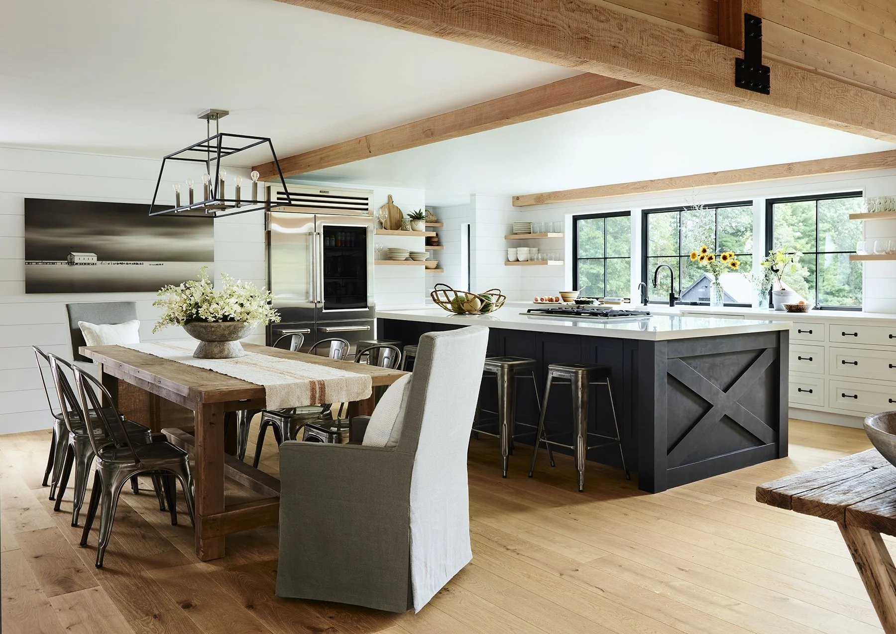 Modern kitchen with white walls, large windows, wooden beams, black kitchen island, dining table with chairs, and decorative items.