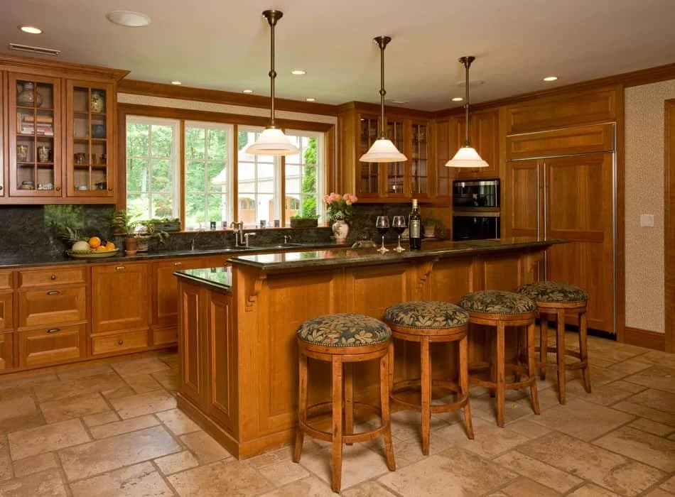 Kitchen with wooden cabinets, a large island with four upholstered bar stools, pendant lights, a window view of greenery, and a black countertop with decorative items and a fruit bowl.