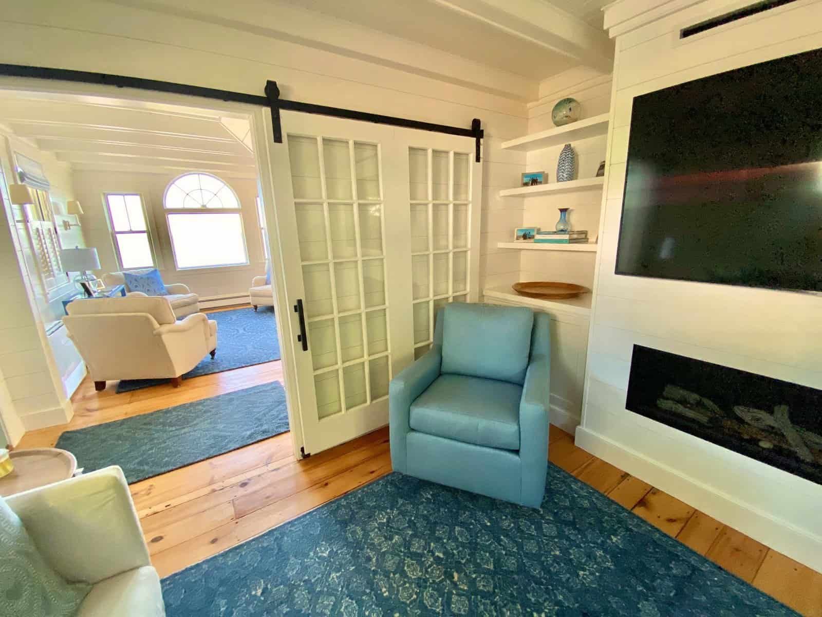 Living room with light-colored walls, a blue armchair, a fireplace, built-in shelves, and a large flat-screen TV, with a glimpse of another sitting area through a doorway.
