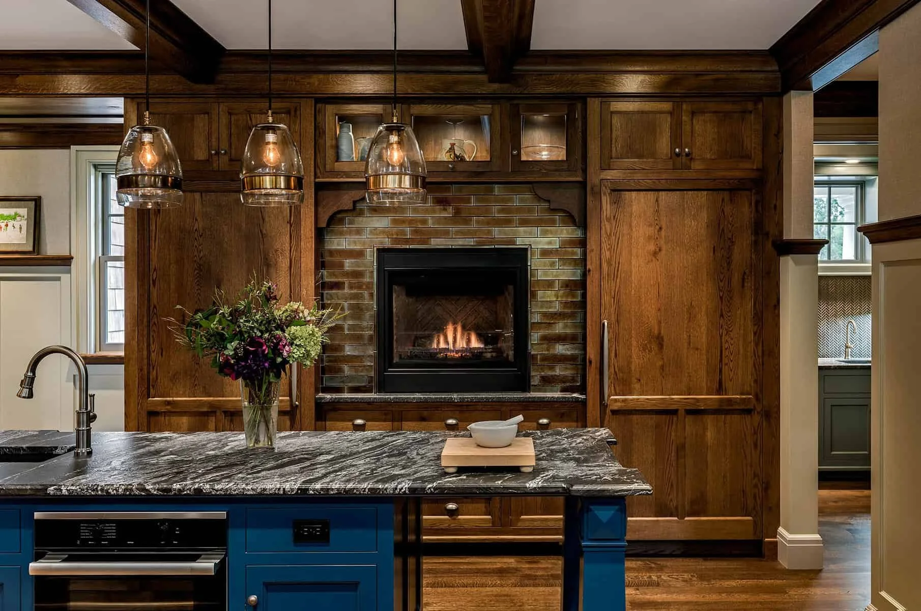 Kitchen with wooden cabinets, a fireplace, and a kitchen island with a black and white marble countertop. A bouquet of flowers is on the island, and three hanging pendant lights are above it. There are windows in the background.
