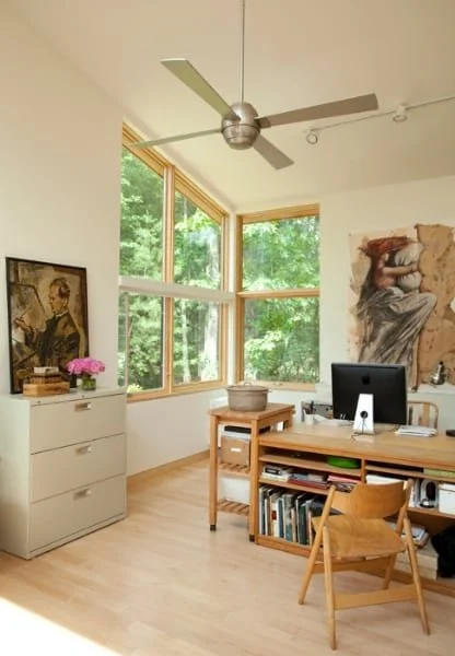 Home office with large windows, a ceiling fan, a desk with a computer, books, and art on the walls.