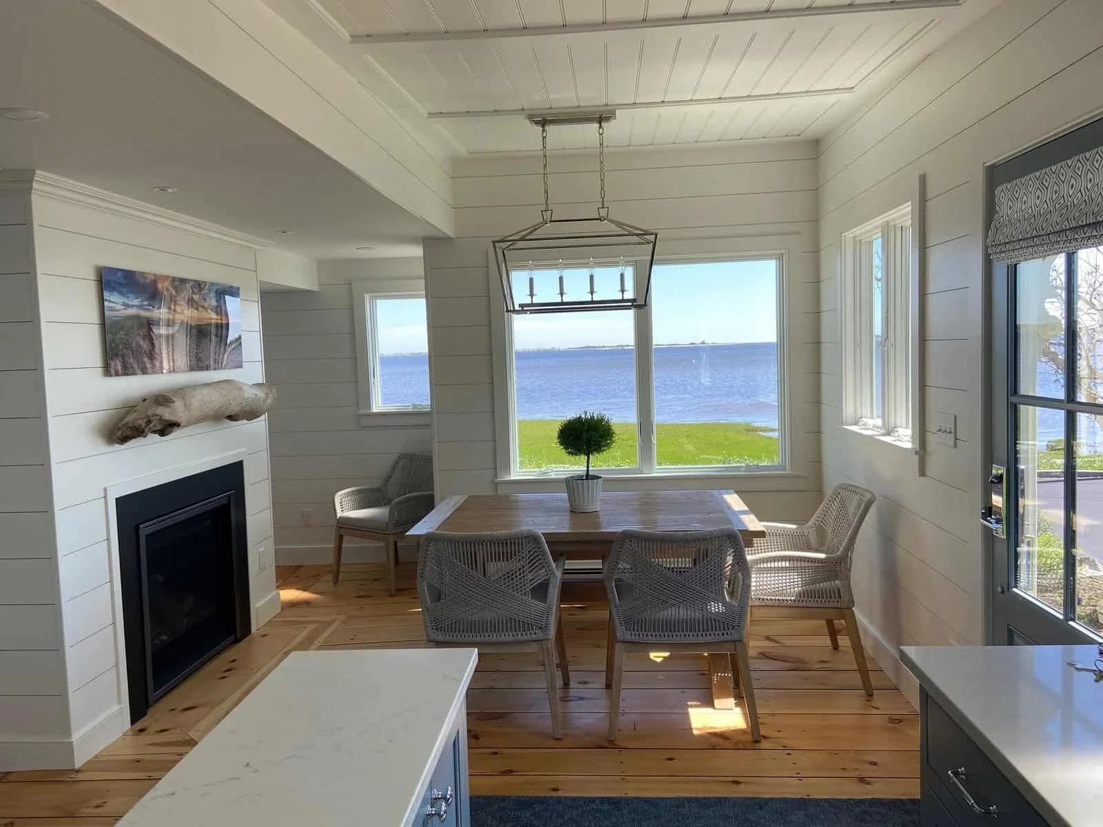 Bright dining room with white shiplap walls and large windows overlooking ocean view, wooden flooring, a rectangular dining table with six wicker chairs, centerpiece plant, and fireplace