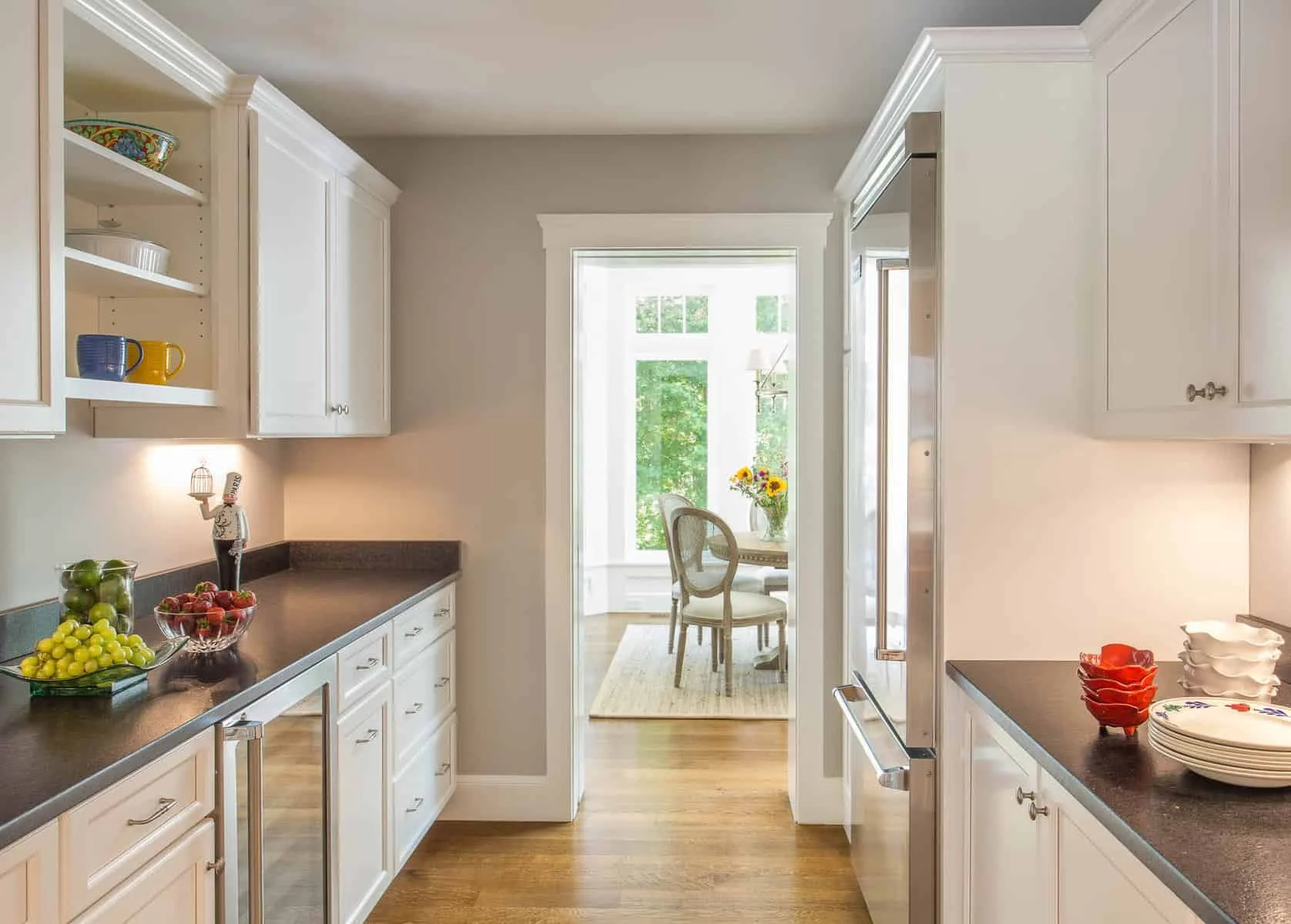 A bright kitchen with white cabinets, dark countertops, and a view of a sunlit dining area with a small table and chairs near a large window with greenery outside.