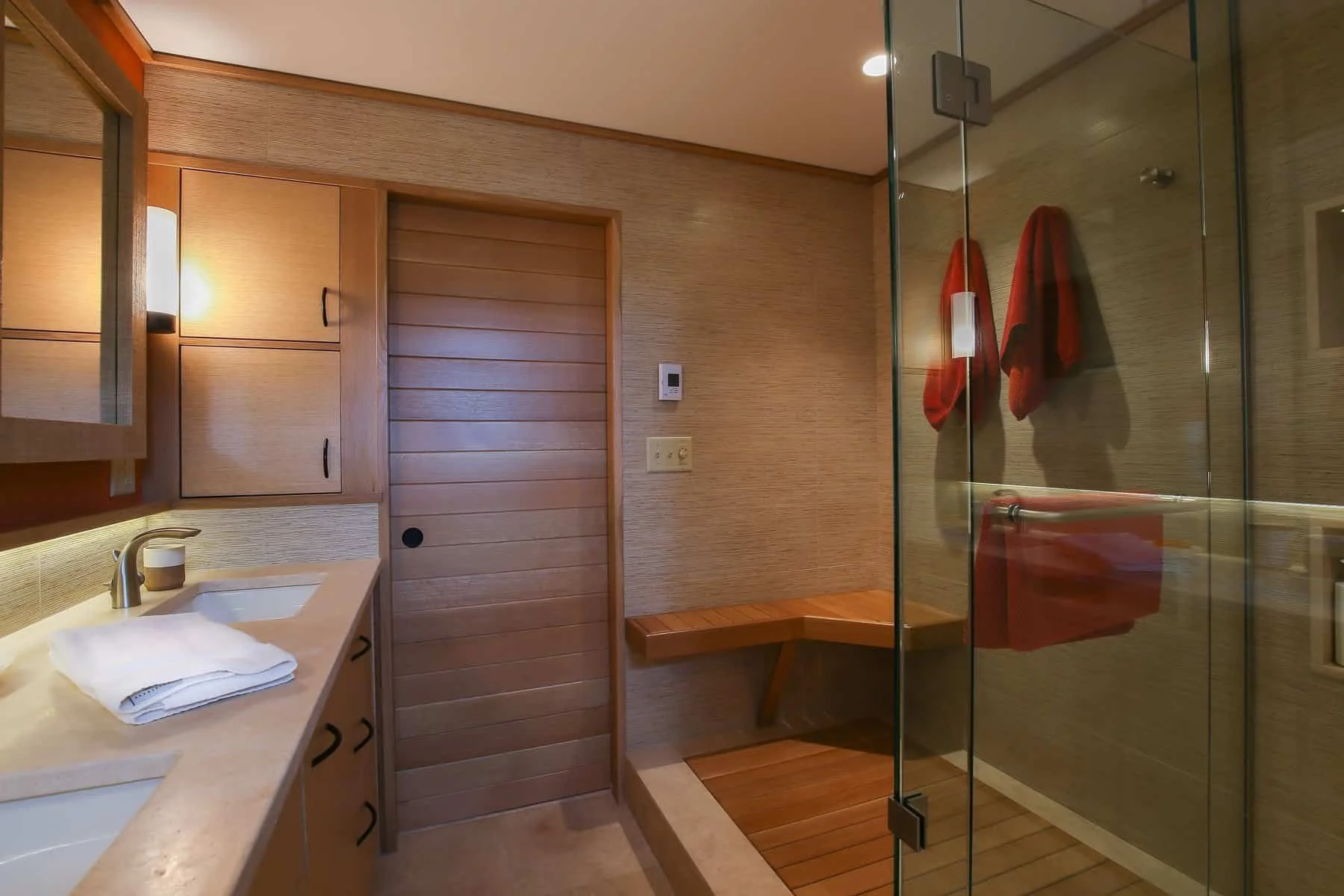 A modern bathroom with wooden cabinets, a beige countertop with a white sink and towels, a wooden bench, a shower with glass doors, and a red towel hanging on the wall.