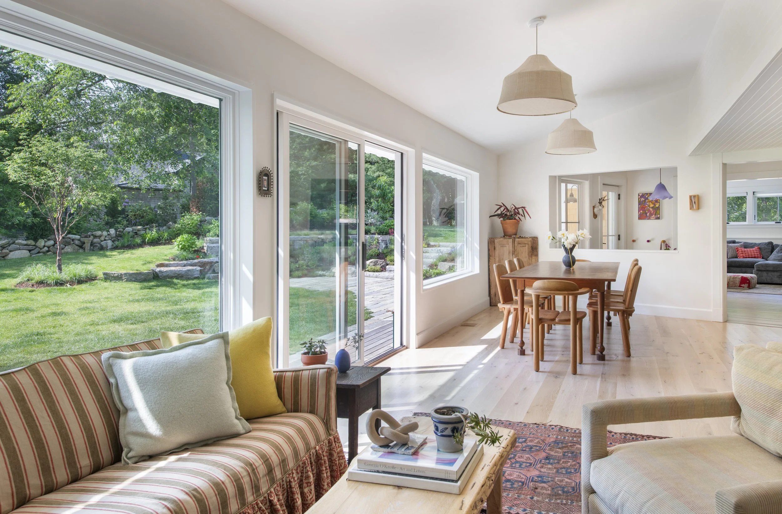 Living room with large sliding glass doors opening to a lush green backyard, featuring patio, trees, and garden. Interior includes patterned sofas with pillows, a wooden dining table with chairs, hanging pendant lights, potted plants, and artwork on 