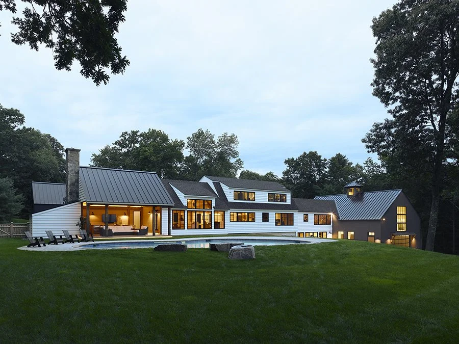 Modern house with large windows, outdoor seating area, and a swimming pool in a lush backyard during dusk.
