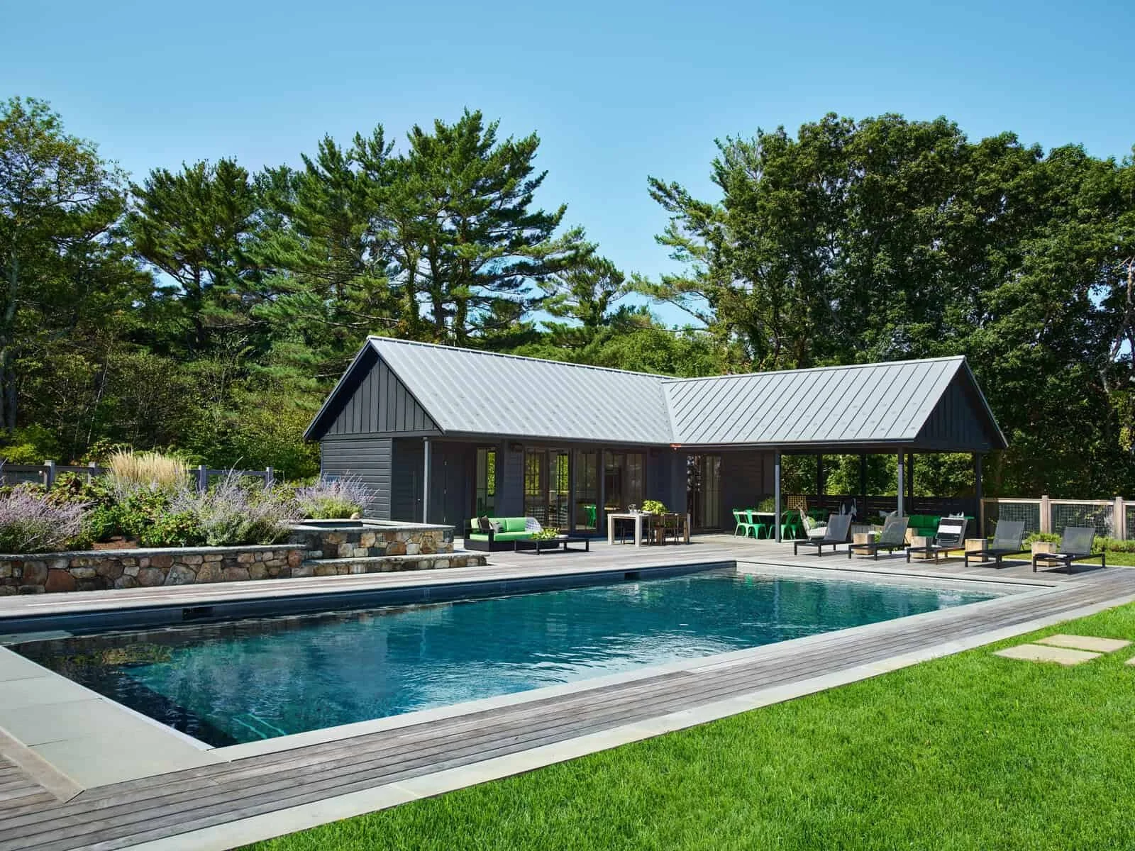 A backyard with a swimming pool, patio furniture, a small modern house or pool house, and green trees in the background under a clear blue sky.