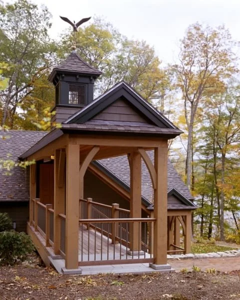 A wooden deck with railings attached to a house with grey shingles, surrounded by trees in fall foliage.