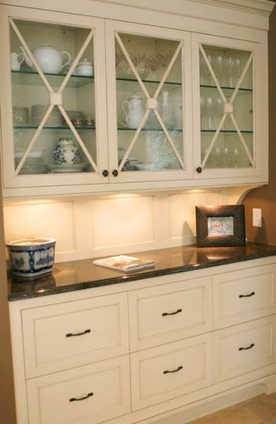 A white kitchen cabinet with glass doors displaying dishes and teapots, with a dark countertop, a framed photo, a decorative bowl, and a book on the counter.