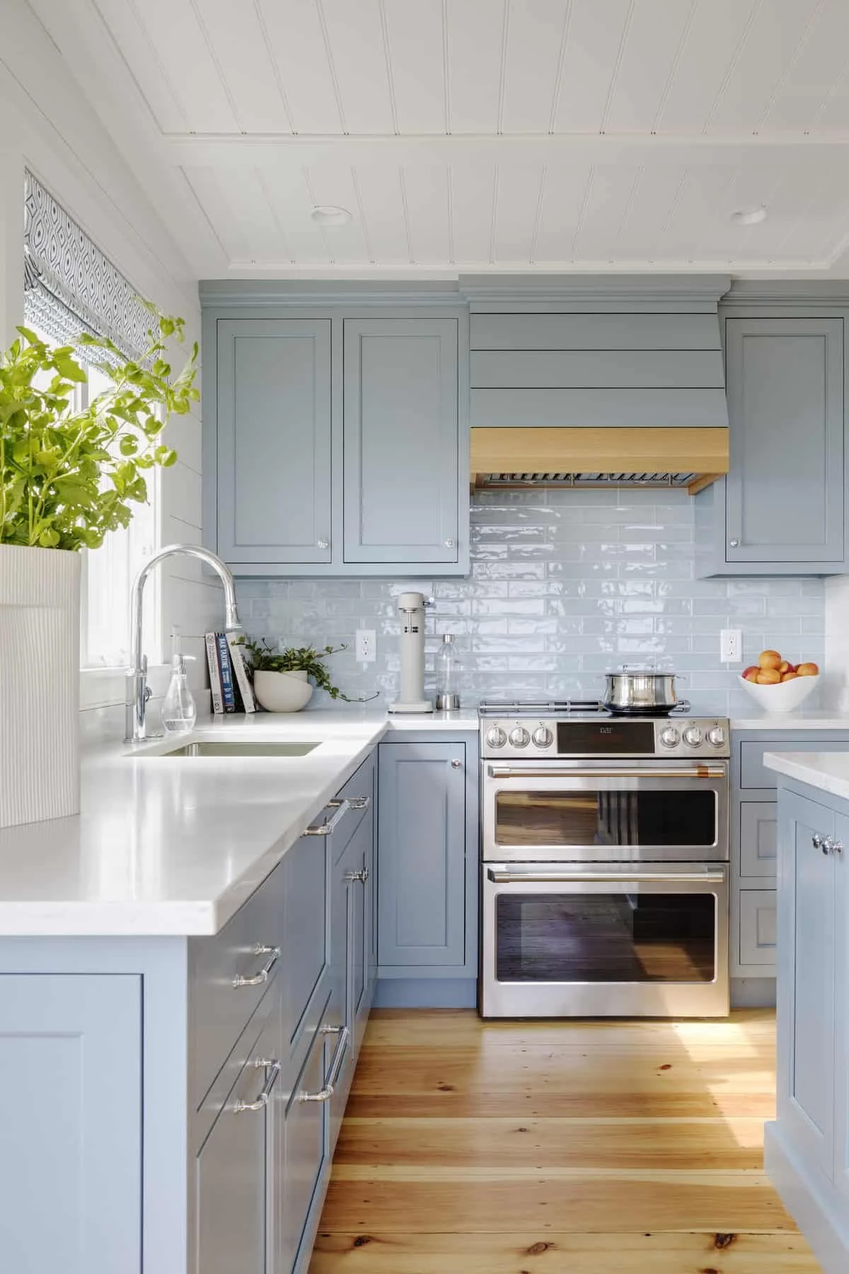 A modern kitchen with light blue cabinets, white countertops, a stainless steel double oven, a white backsplash, and wood flooring. There are green plants, books, and a bowl of peaches on the counter.