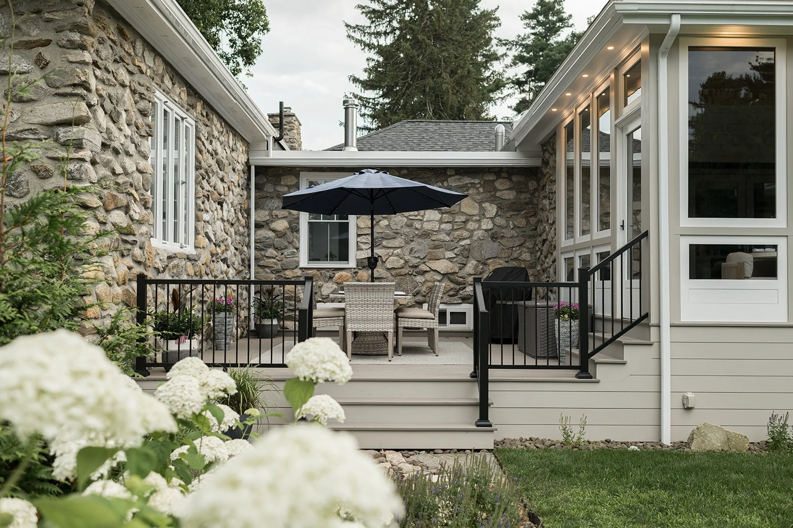 Backyard patio with stone wall, outdoor dining table, chairs, black umbrella, potted plants, grill, and enclosed by black railing, attached to house with large windows and steps leading down to garden.
