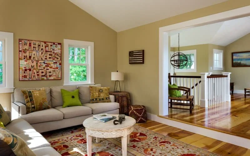 Living room with beige walls, white sofa, colorful pillows, a round coffee table, and artwork on the wall, adjacent to a second sitting area visible through an open doorway.