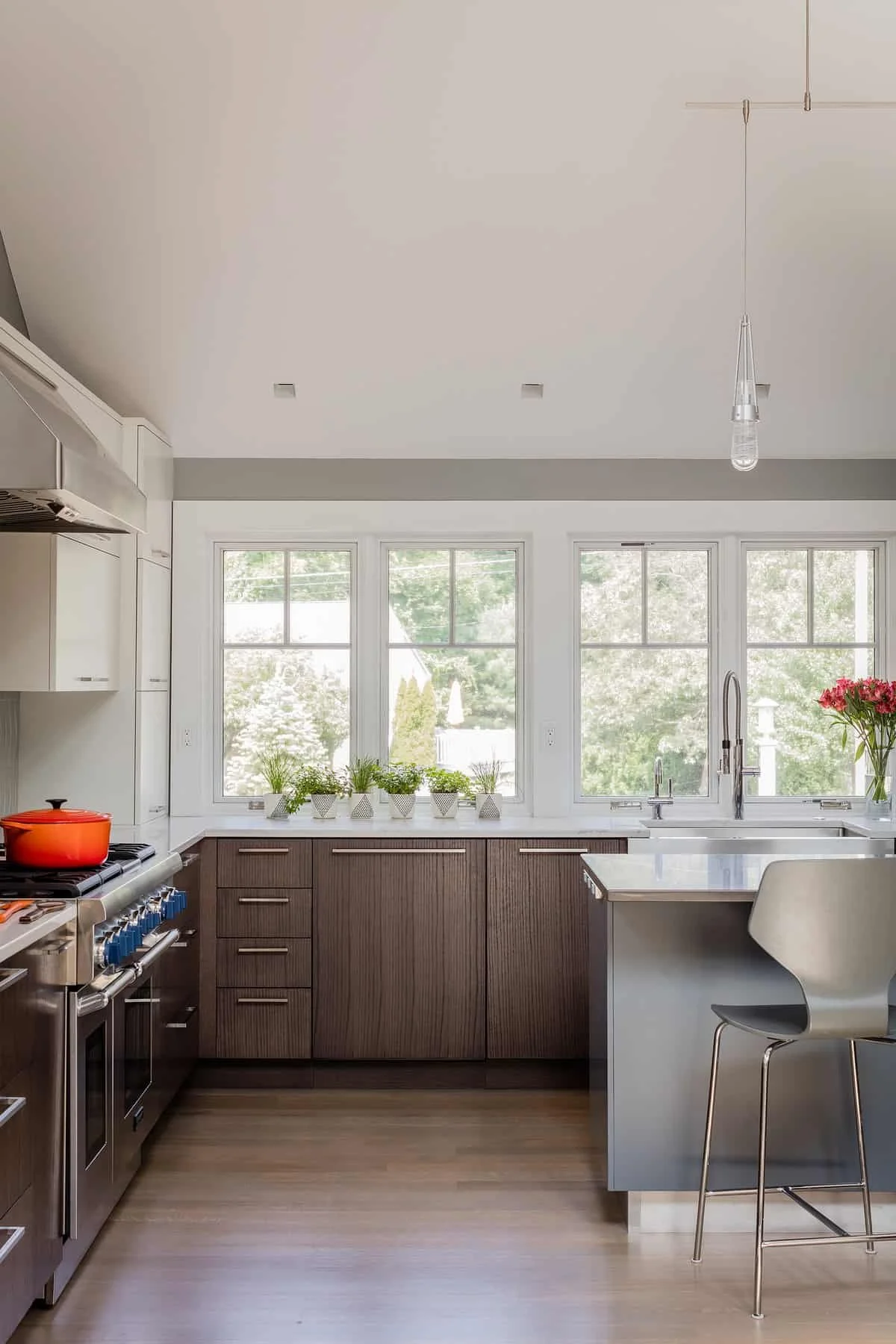 Modern kitchen with white upper cabinets, dark wood lower cabinets, a marble countertop, large windows with potted plants, and a white chair with a metal frame at a kitchen island.