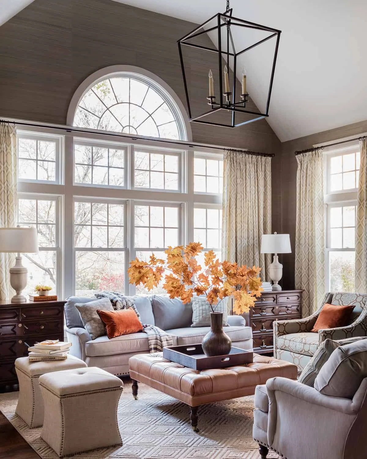 A cozy living room with large windows, beige curtains, a white sofa with orange pillows, a matching armchair, and a coffee table with a vase of orange autumn leaves.