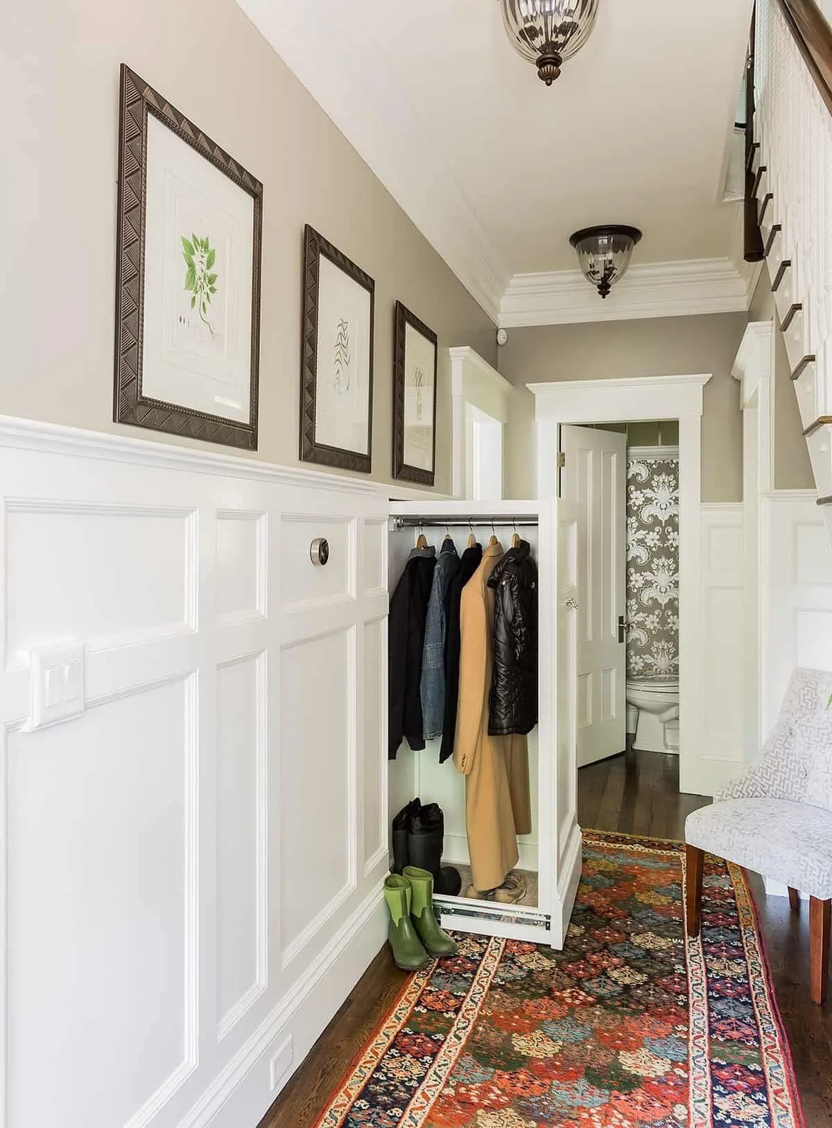 Entryway with white wainscoting, framed artwork on the wall, a coat rack with jackets and shoes, a patterned rug, and a doorway leading to a bathroom with floral wallpaper.