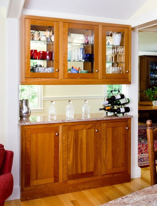 A wooden china cabinet with glass doors on top, filled with glassware and decorative items, and solid wooden doors on the bottom, situated against a wall in a dining area.