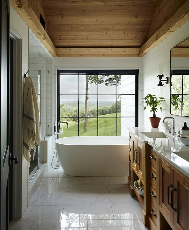 Modern bathroom with freestanding bathtub, large window showing an outdoor view of trees and grassy landscape, wooden ceiling, white tiled floor, and wooden cabinetry with a potted plant on the countertop.