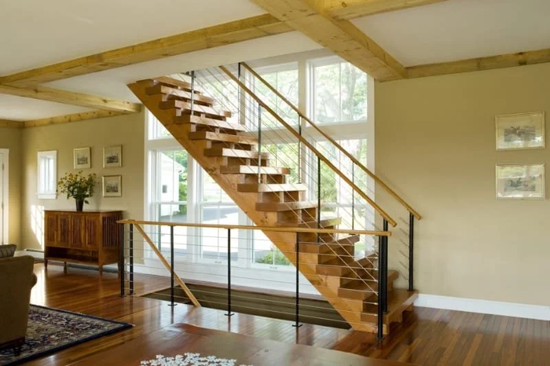Interior view of a modern living room featuring a wooden staircase with a metal railing, large windows allowing natural light, a wooden sideboard with a vase of flowers, framed artwork on the walls, and hardwood floors.