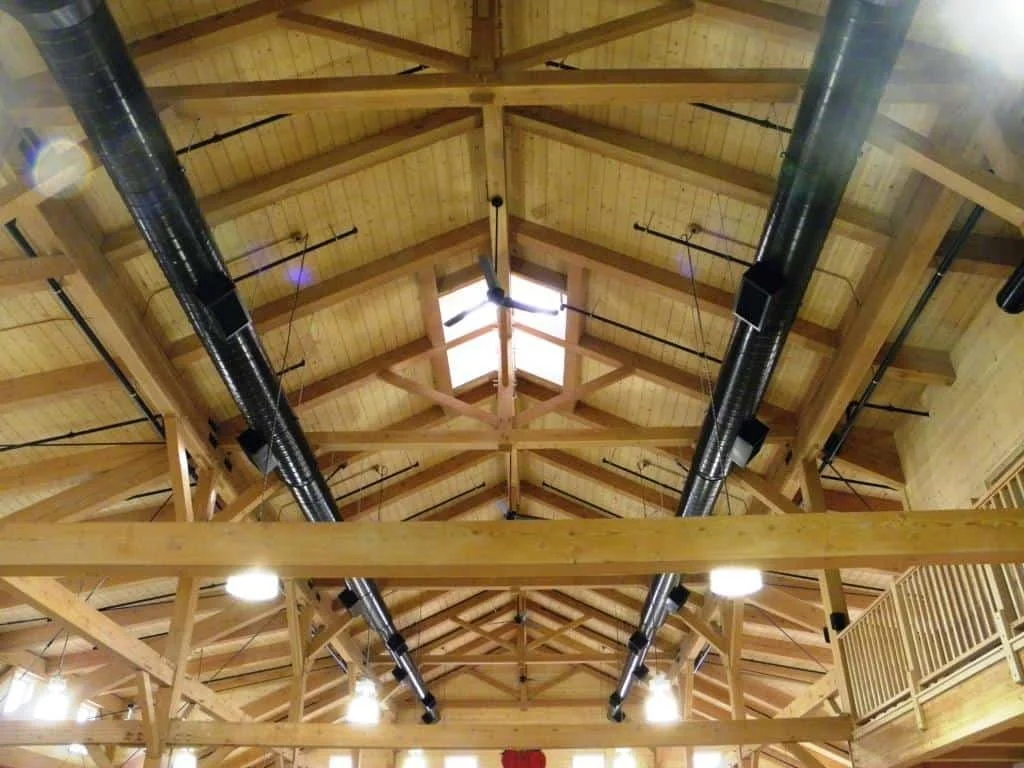 Interior view of a wooden ceiling with exposed beams, black ventilation ducts, and ceiling lights.