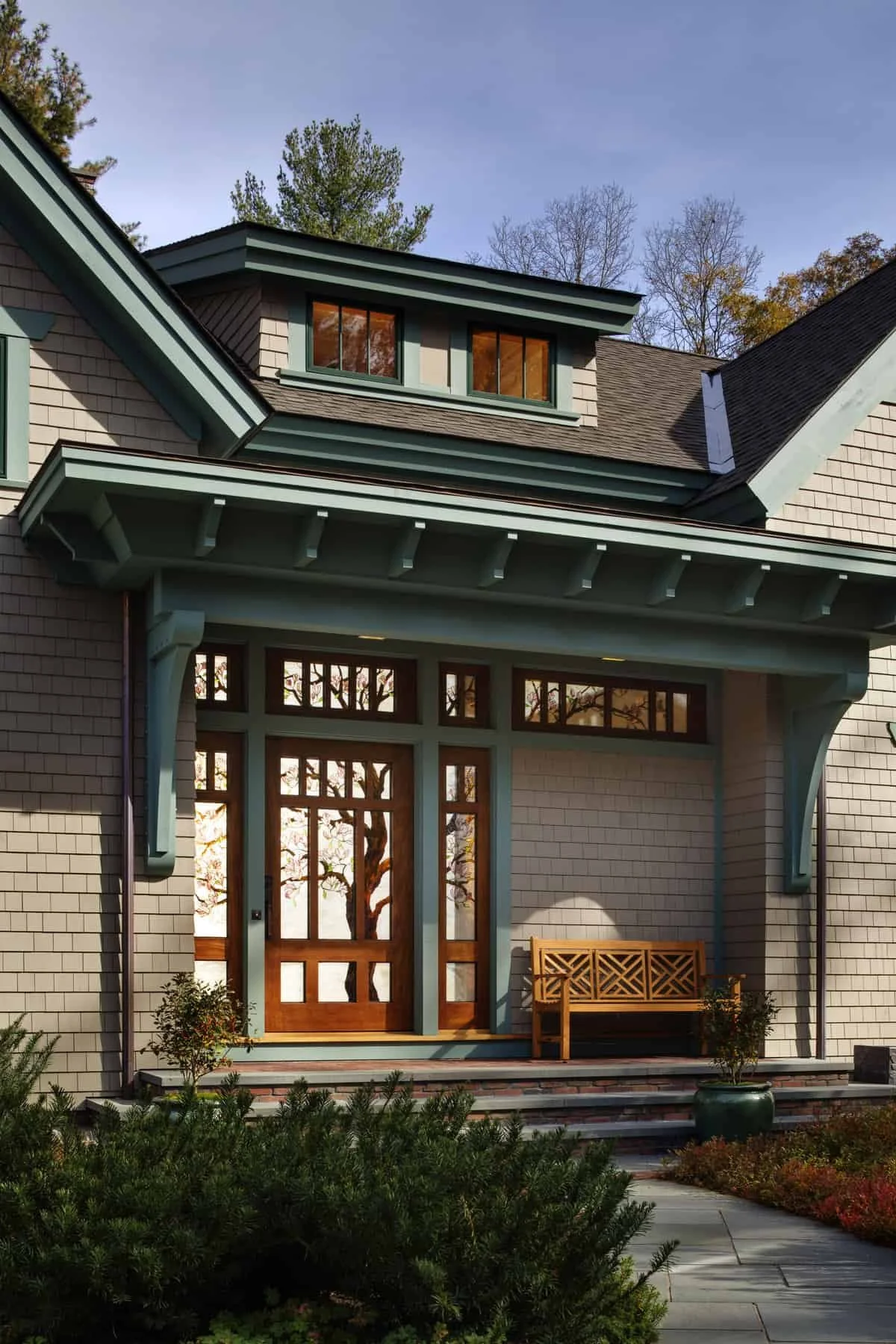 Front porch of a house with a wooden door, decorative glass, and a wooden bench. The house has beige brick siding with teal trim and a large overhang. There are small trees in pots and landscaped bushes surrounding the porch, with a clear sky and tre