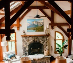 Living room with stone fireplace, wooden beams, large windows, and a ceiling fan.