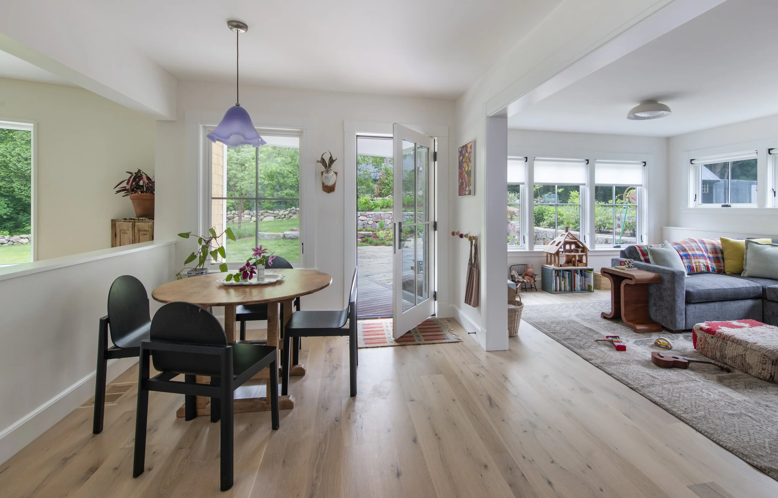 Open-concept living and dining room with large windows showcasing a garden, wooden floors, a dining table with black chairs, and a gray sofa with colorful pillows.