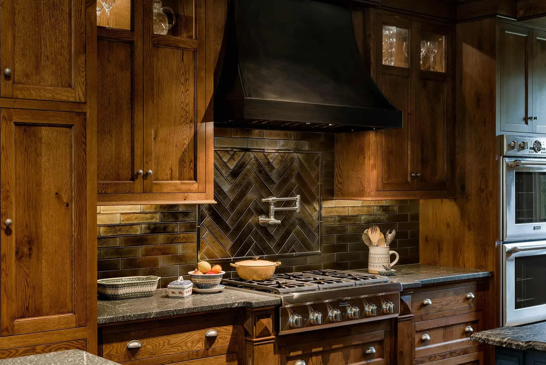 A kitchen with wooden cabinets, a stove with oven, a black range hood, tiled backsplash with a herringbone pattern, a sink with a dish scrubber, and various kitchen items on the countertop.