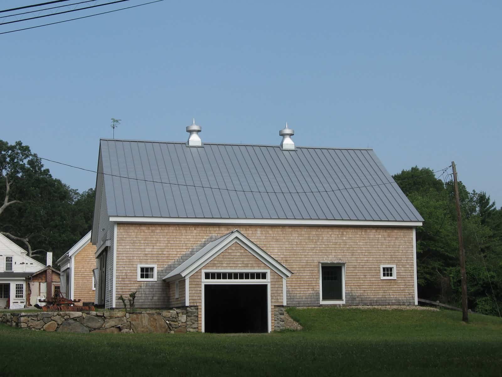 A large barn with a gabled metal roof and wooden shingle siding, featuring a garage door and small windows, surrounded by a grassy lawn and trees in the background.