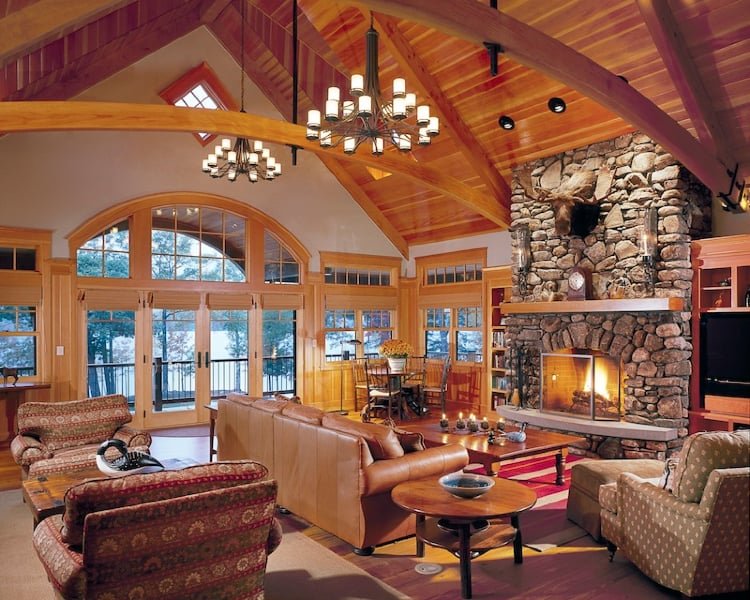 Cozy living room with a large stone fireplace, high wood-paneled ceiling with exposed beams, multiple large windows, and a mix of vintage and modern furniture. There are chandeliers hanging from the ceiling and a fire burning in the fireplace.