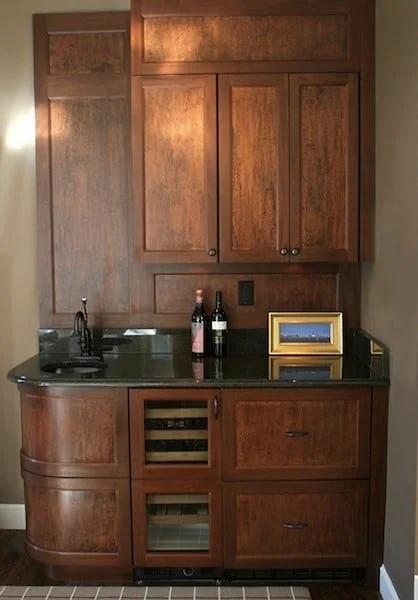 Wooden kitchen cabinet with a small sink, two wine bottles, and a framed picture on the counter.