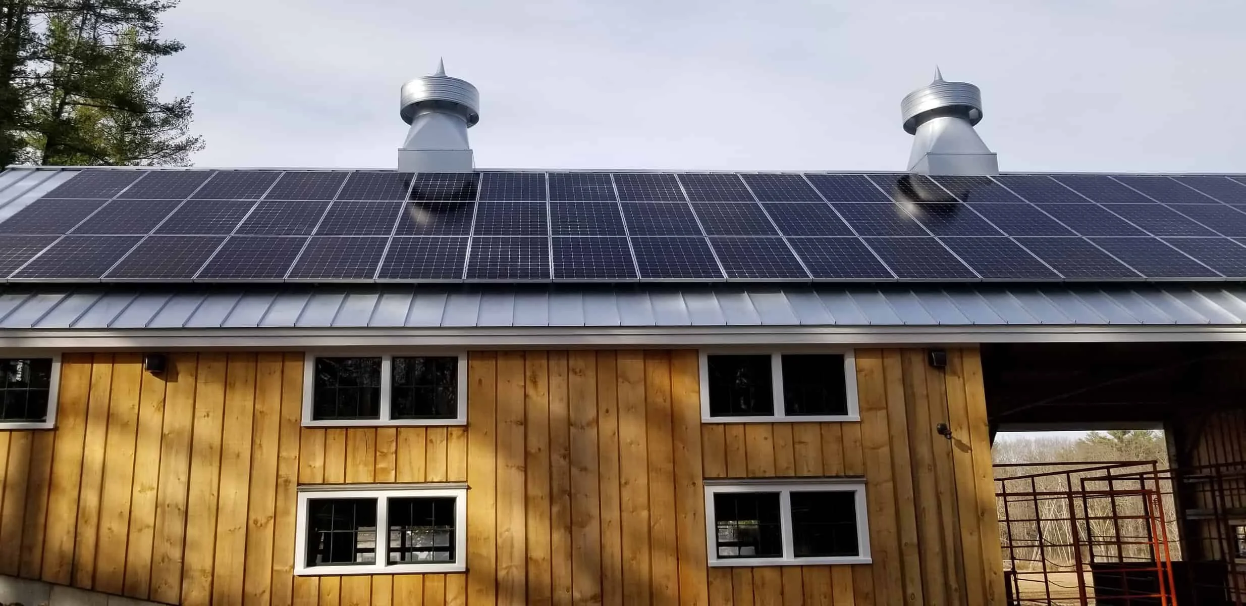 A house with a wooden exterior and a metal roof covered with solar panels. Two vents are visible on the roof, and in the background, there are tall trees and a cloudy sky.