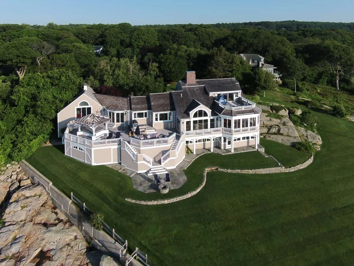 Large house with multiple decks and balconies, surrounded by a landscaped yard and dense trees, positioned on a hillside.