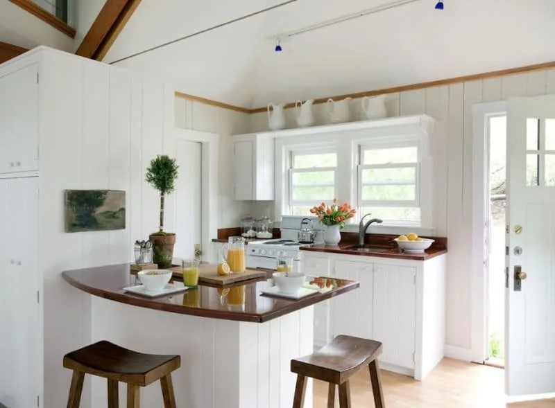 Bright white kitchen with wooden accents, a small island with stools, a window above the sink, breakfast items on the counter, and a door to the outside.