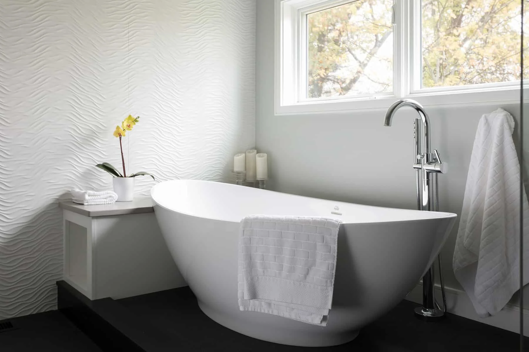 Modern bathroom with a freestanding bathtub near a window, decorated with a potted orchid, candles, and white towels.