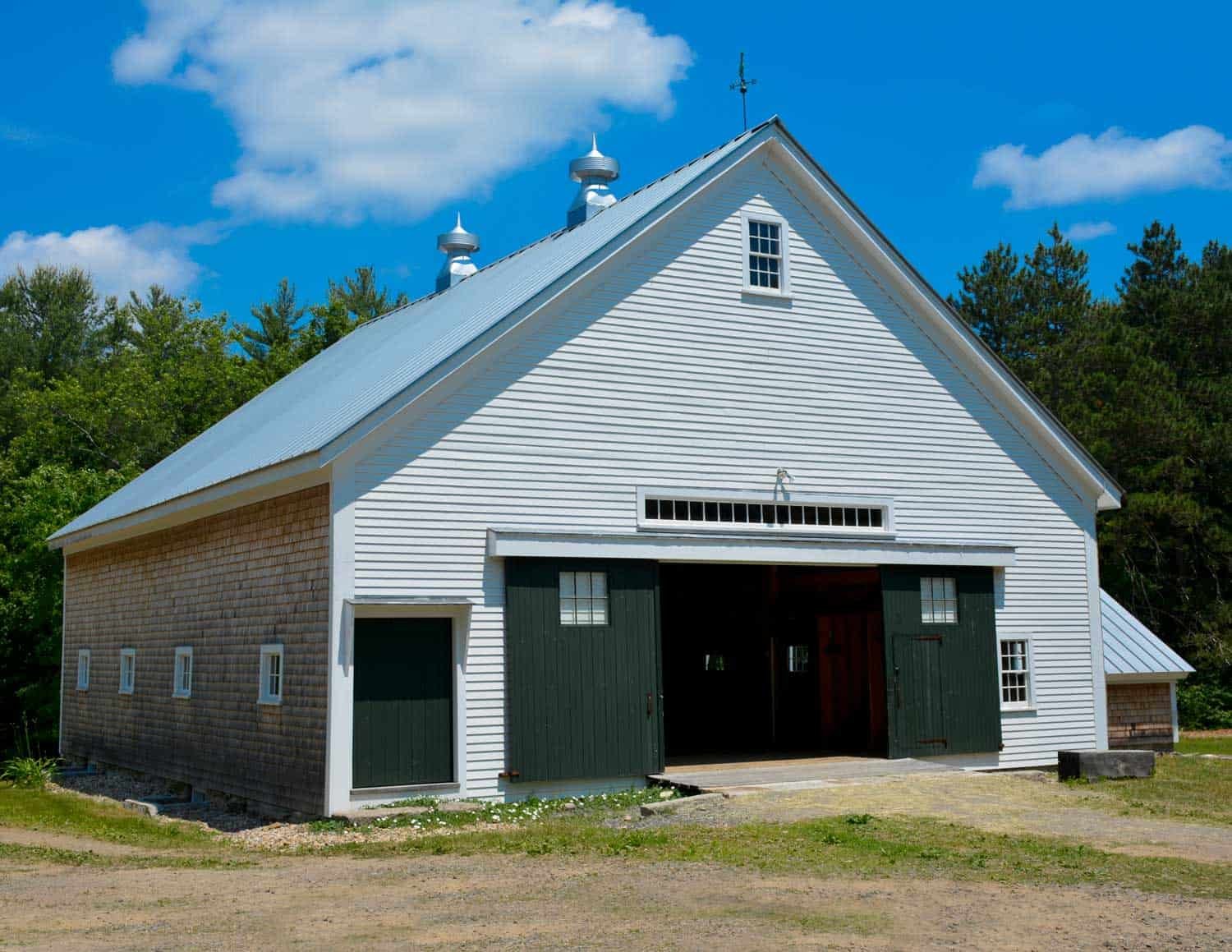 White barn with green doors and shingles, set against a backdrop of trees and a blue sky with clouds.