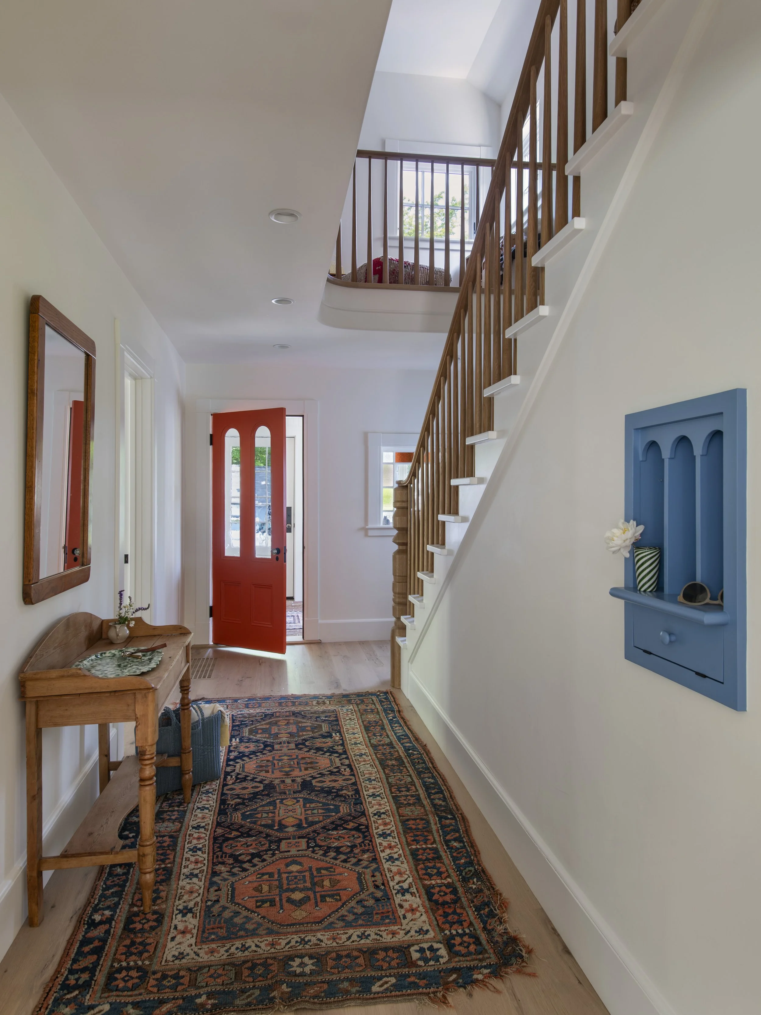 Entrance hallway of a house with a red front door, wooden staircase on the right, and decorative rug on the floor. A blue shelf with small decorative items is mounted on the wall on the right side.