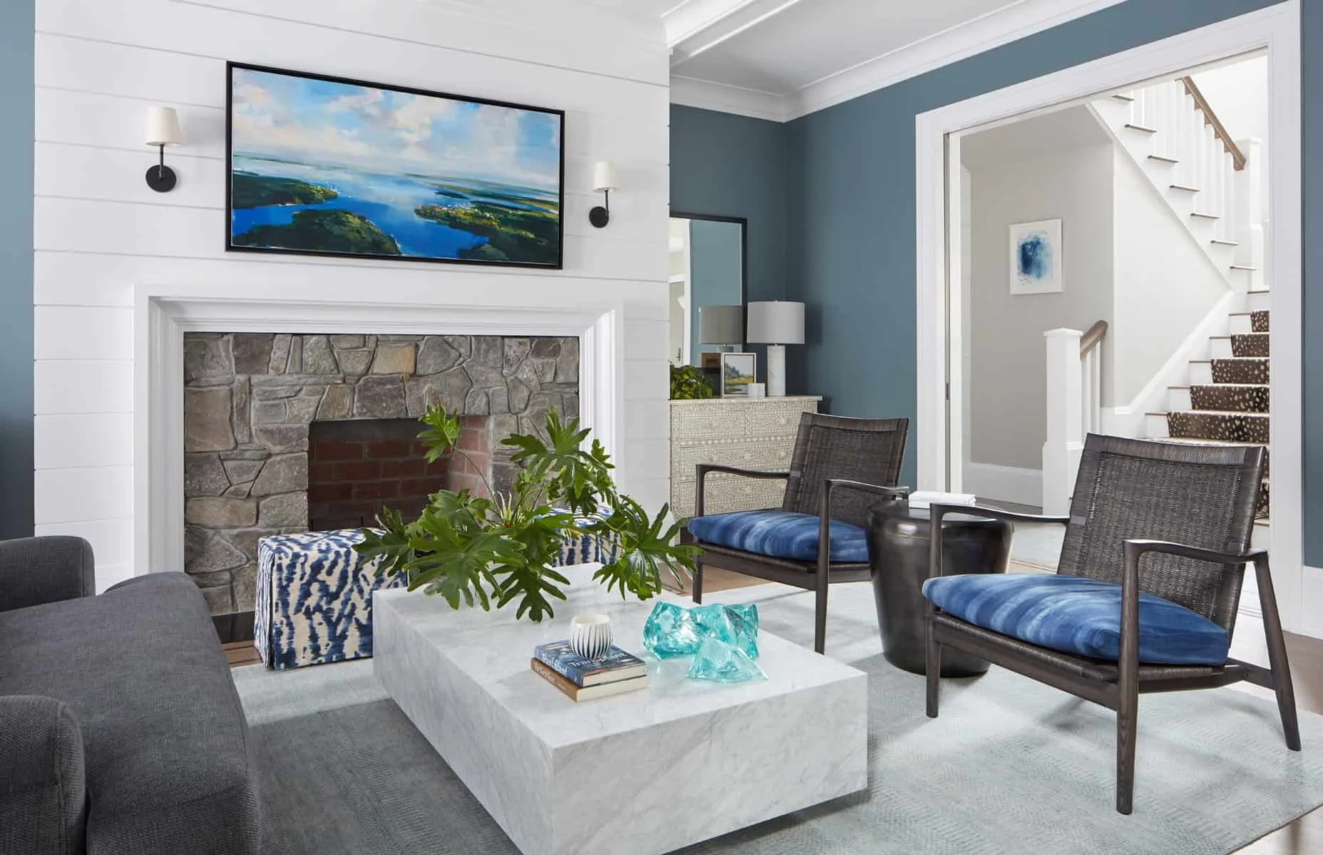 Living room with a stone fireplace, a wall-mounted TV, a white coffee table with decorative objects, and blue cushion armchairs, featuring a staircase visible through a doorway.