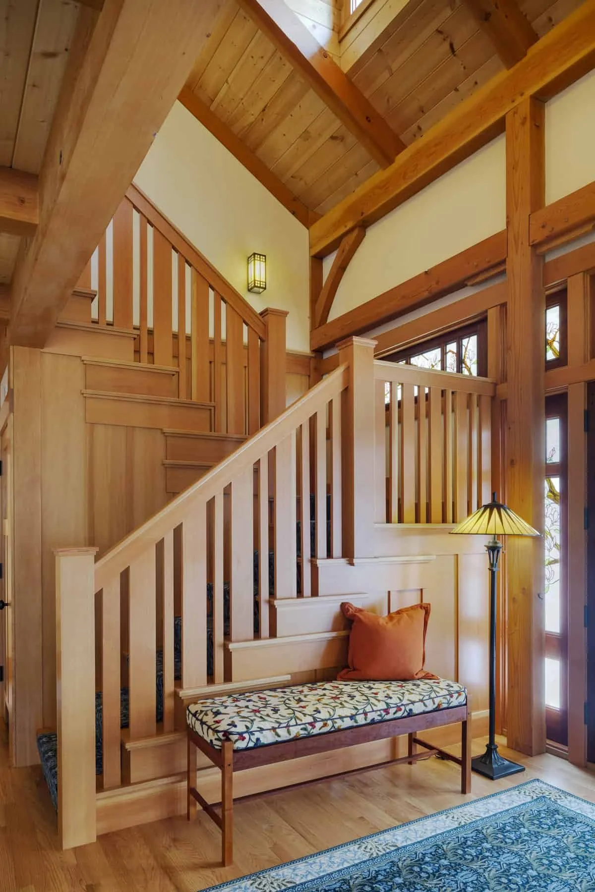 A wooden staircase in a home with high ceilings, a decorative bench with floral upholstery and an orange pillow, a standing lamp with a yellow lampshade, and a window with wooden trim in the background.