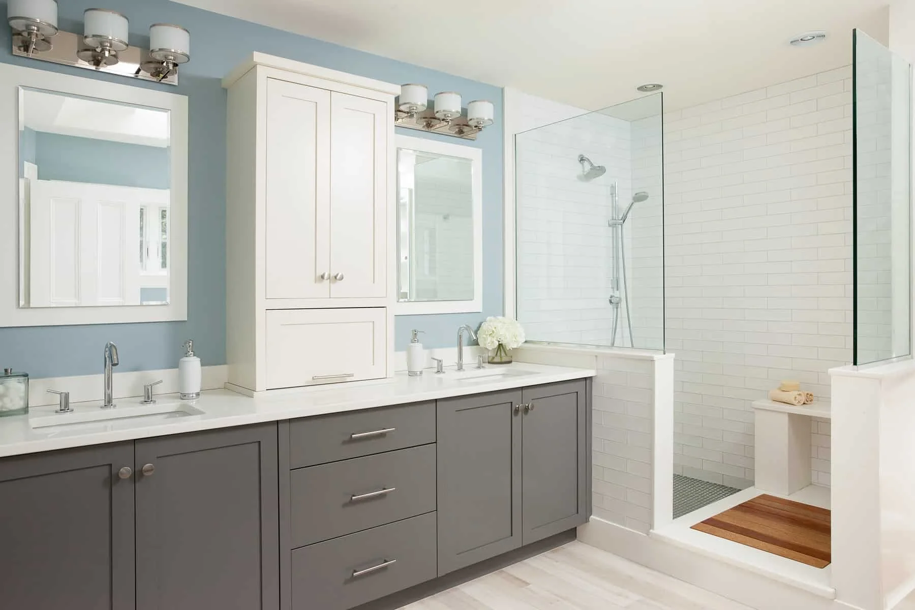 A modern bathroom with a double sink vanity, light blue wall, white cabinets with mirrors, and a walk-in shower with white subway tiles and a glass partition.