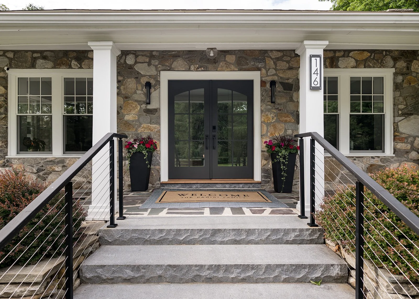 Front porch of a house with stone exterior, black front door with glass panels, two black planters with pink and red flowers on the sides, and two windows with white frames. There is a welcome mat with the word 'Welcome' and house number 146 on a whi