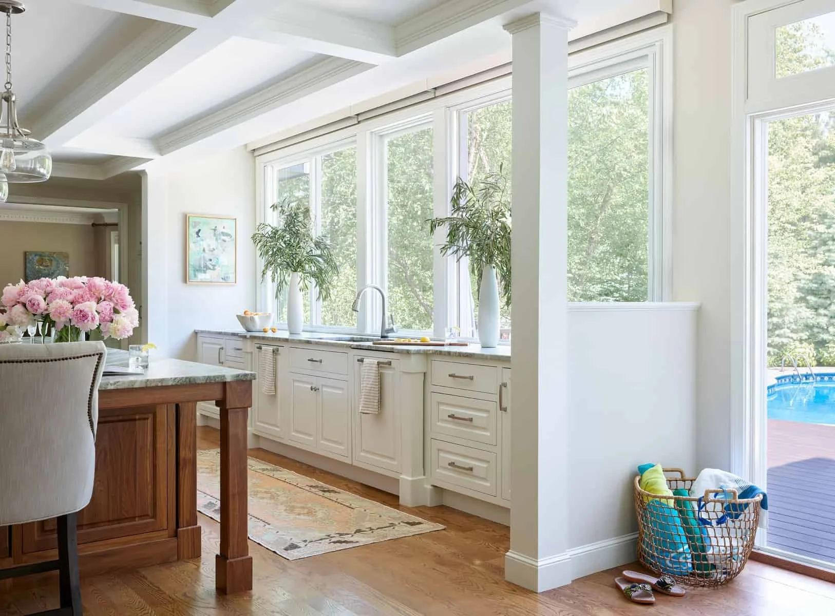Bright kitchen with large windows, white cabinets, a marble countertop, and green plants, with a view of a backyard pool outside.