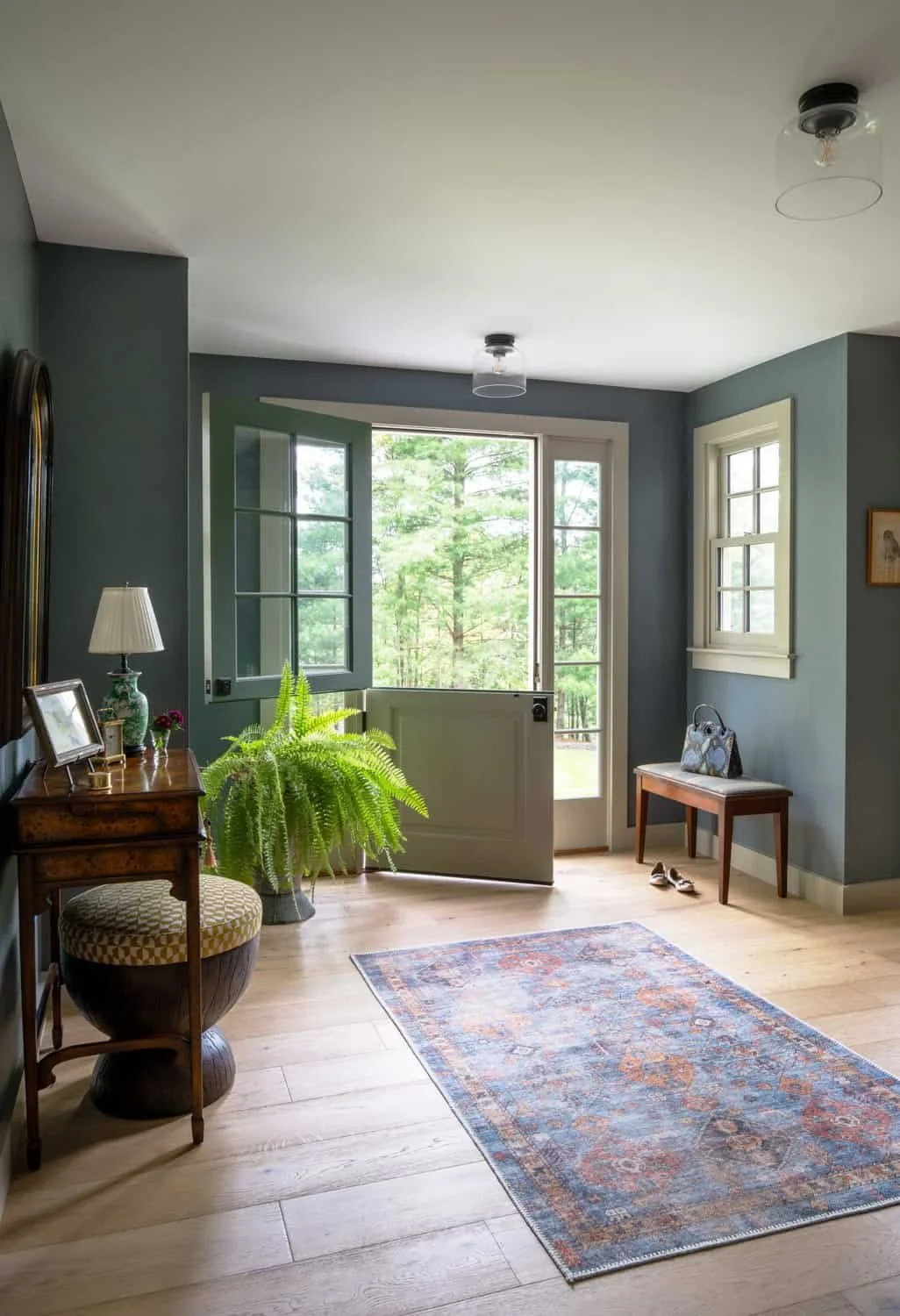 Bright living room with an open door leading outside to a green forest, a wooden side table with a lamp and picture frame, a large potted fern, a small wooden bench with a bag, and a pair of shoes on the floor. Light blue walls and a wooden floor wit
