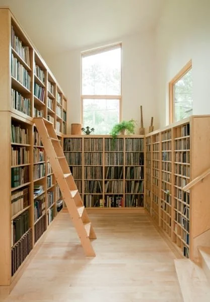 A cozy home library with tall wooden bookshelves filled with books, a ladder leaning against the shelves, and large windows letting in natural light.