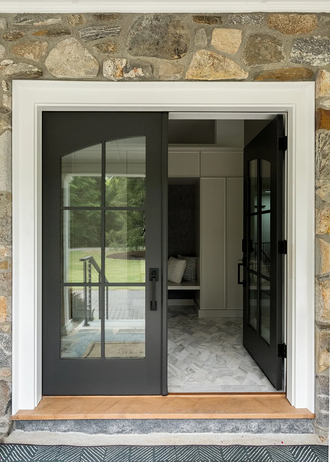 Exterior view of a house showing a glass double door with black frames, leading into a modern interior with white cabinets, a bench with pillows, and marble flooring. The door is surrounded by a stone wall.